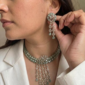 Op de afbeelding: Elegant zilverkleurig sieradenset bestaande uit een ketting en oorbellen. De ketting heeft een dubbelstrengs ontwerp met ovale groene stenen en een centrale hanger met bungelende accenten. De oorbellen weerspiegelen het ontwerp met een cirkelvormige bovenkant en trapsgewijze druppels.
