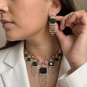 May include: A matching jewelry set featuring a necklace and earrings. The necklace has a pink chain with emerald green rectangular stones and dangling diamond-like accents. The earrings have a similar design, with a green stone and dangling accents. The jewelry is set against a white blazer.