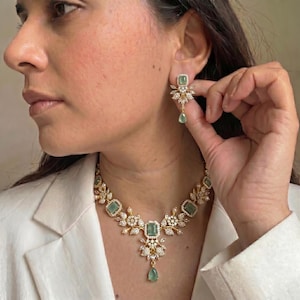 May include: A gold-toned necklace and earring set featuring emerald-green rectangular and teardrop-shaped gemstones. The necklace and earrings have a floral design with clear, sparkling accents. The jewelry is worn with a white blazer.