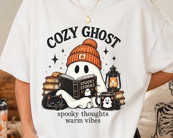 May include: A white t-shirt featuring a graphic of a ghost wearing an orange beanie, reading a book titled Spooky Stories. The design includes a black cat, a stack of books, a lantern, and a mug, with the text Cozy Ghost, spooky thoughts, warm vibes.