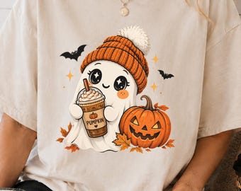 May include: A beige short-sleeve t-shirt featuring a graphic of a white ghost wearing an orange knit beanie, holding a coffee cup labeled PUMPKIN, and standing next to a jack-o'-lantern with orange autumn leaves, small bats, and stars.