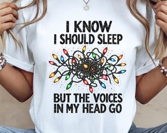 May include: A white short-sleeve t-shirt featuring a graphic of tangled, colorful Christmas lights in the center. The text reads: I KNOW I SHOULD SLEEP BUT THE VOICES IN MY HEAD GO. The lettering is black with a distressed, textured appearance.