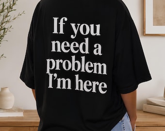 May include: A black short-sleeved t-shirt featuring white text on the back that reads, "If you need a problem I'm here." The shirt has a crew neckline and a loose, relaxed fit, paired with light blue denim jeans with visible back pockets.