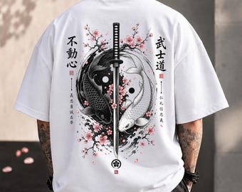 May include: A white short-sleeved t-shirt featuring a back graphic of a vertical katana sword dividing a black and white yin-yang koi fish design, surrounded by pink cherry blossoms and Japanese kanji text on both sides.