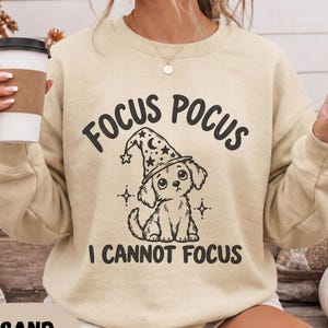 May include: A sand-coloured crewneck sweatshirt featuring a black graphic of a puppy wearing a wizard hat. The text above the puppy reads FOCUS POCUS and the text below reads I CANNOT FOCUS. The design includes small star and sparkle accents.
