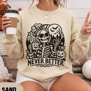May include: A sand-colored crewneck sweatshirt featuring a black graphic print of a skeleton wearing sunglasses and a headband, holding a jack-o'-lantern mug. The design includes bats, trees, and the text NEVER BETTER in bold letters.