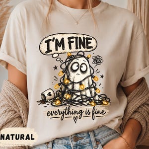 May include: A natural-coloured t-shirt featuring a graphic of a ghost tangled in yellow string lights with a spilled coffee mug. Above the ghost, a speech bubble contains the text "I'M FINE," and below, the text "everything is fine" with a heart.