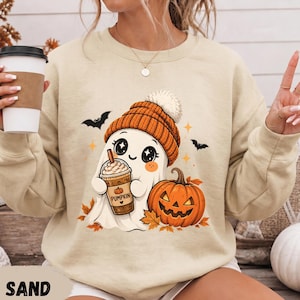 Op de afbeelding: Een zandkleurige sweater met ronde hals en een afbeelding van een wit spookje met een oranje gebreide muts, dat een koffiebeker met het opschrift PUMPKIN vasthoudt, naast een pompoen en herfstbladeren, met kleine zwarte vleermuizen op de achtergrond.