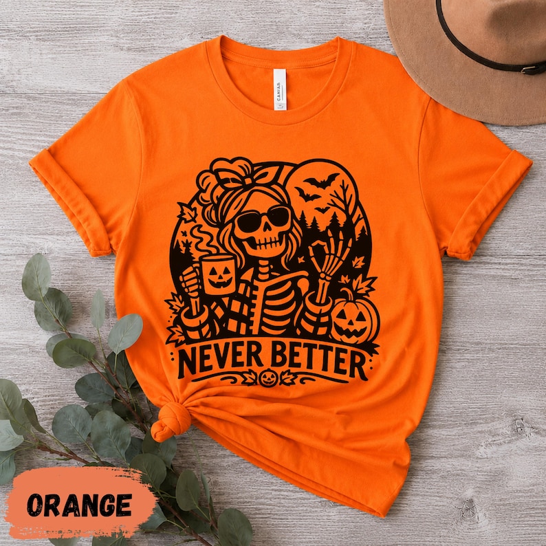 May include: A bright orange short-sleeve t-shirt featuring a black graphic print of a skeleton wearing sunglasses, holding a jack-o'-lantern mug, and making an okay hand gesture. The design includes bats, trees, and the text NEVER BETTER.