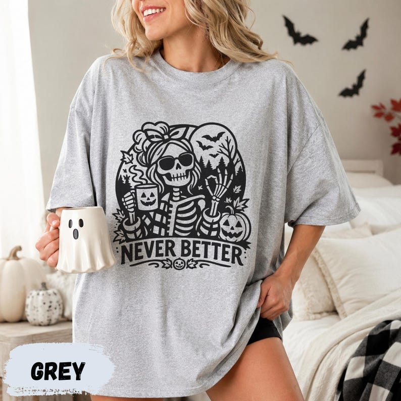May include: A light grey t-shirt featuring a black graphic of a skeleton wearing sunglasses and a hair bow, holding a jack-o'-lantern mug. The design includes bats, trees, and a pumpkin with the text "NEVER BETTER" printed below.