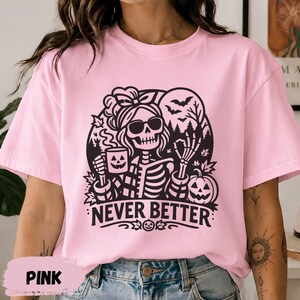 May include: A light pink t-shirt featuring a black graphic of a skeleton wearing sunglasses and a hair bow. The skeleton holds a jack-o'-lantern mug and makes an okay hand gesture. The text NEVER BETTER is printed below the design.