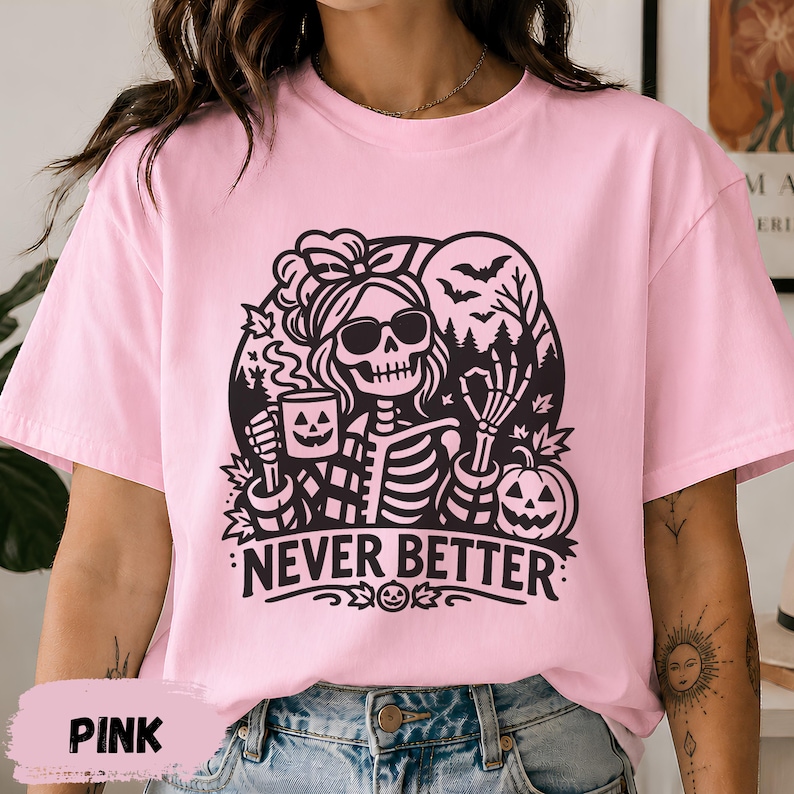 May include: A light pink t-shirt featuring a black graphic of a skeleton wearing sunglasses and a hair bow. The skeleton holds a jack-o'-lantern mug and makes an okay hand gesture. The text NEVER BETTER is printed below the design.