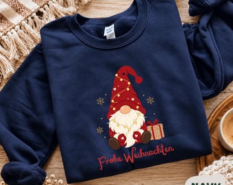 May include: A navy blue crewneck sweatshirt featuring a graphic of a gnome wearing a red hat with string lights and hearts. The gnome sits next to a gift box, surrounded by gold snowflakes. The text "Frohe Weihnachten" is printed in red below.