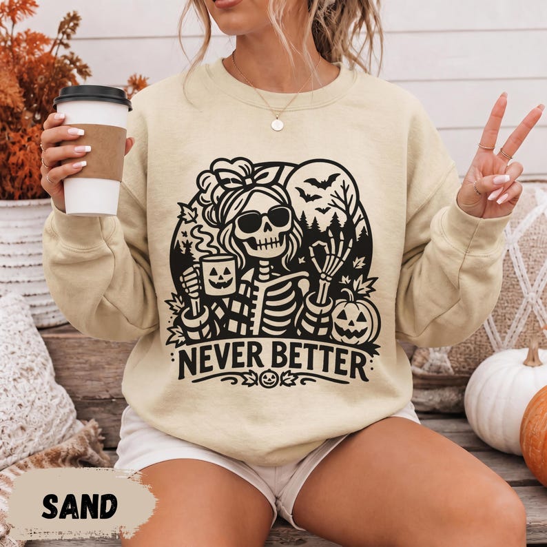 May include: A sand-colored crewneck sweatshirt featuring a black graphic print of a skeleton wearing sunglasses and a headband, holding a jack-o'-lantern mug. The design includes bats, trees, and the text NEVER BETTER in bold letters.