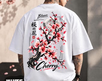 May include: A white short-sleeved t-shirt featuring a graphic on the back of a cherry blossom tree branch with pink flowers, a moon, and the text Blossom, Cherry, and Japanese characters. The shirt has a loose, boxy fit.