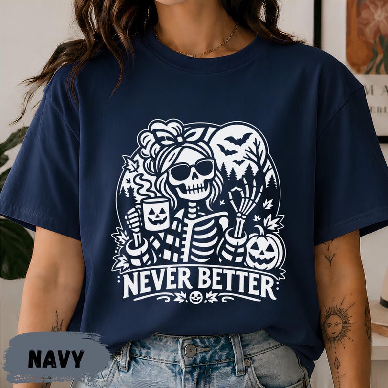 May include: A navy blue t-shirt featuring a white graphic of a skeleton wearing sunglasses and a hair bow, holding a jack-o'-lantern mug. The design includes bats, trees, a pumpkin, and the text NEVER BETTER in bold capital letters.