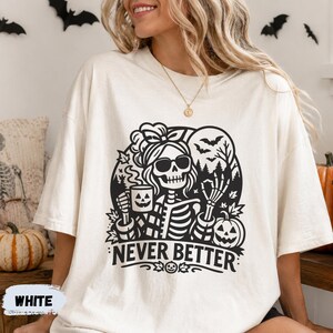 May include: An off-white short-sleeve t-shirt featuring a black graphic print of a skeleton wearing sunglasses and a hair bow, holding a jack-o'-lantern mug. The design includes bats, trees, and the text NEVER BETTER.