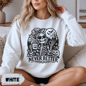 May include: A white long-sleeve sweatshirt featuring a black graphic print of a skeleton wearing sunglasses and a hair bow. The skeleton holds a jack-o'-lantern mug, with a pumpkin, bats, and trees in the background. The text reads NEVER BETTER.