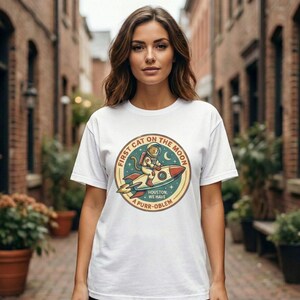 May include: White t-shirt featuring a circular graphic of a cat in an astronaut suit riding a rocket ship. The graphic includes the text "FIRST CAT ON THE MOON" and "HOUSTON, WE HAVE A PURR-OBLEM". The design has a vintage, retro style.