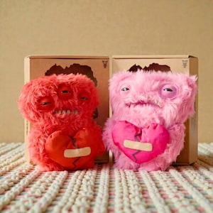 Op de afbeelding: Twee pluche monsterpoppen, één rode en één roze, die elk een gebroken hart vasthouden. De rode pop heeft drie ogen en de roze pop heeft één oog. Beide poppen hebben een grijns en staan voor kartonnen dozen.