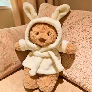 May include: A plush brown teddy bear wearing a white hooded robe with bunny ears. The robe is tied at the waist with a matching belt. The bear is sitting on a pink pillow.