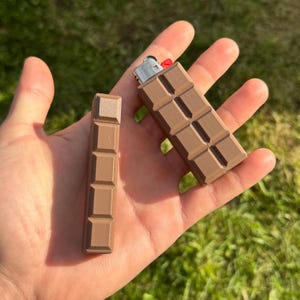 Könnte beinhalten: Zwei braune Feuerzeuge in Form von Schokoladentafeln. Eines ist ein Standardfeuerzeug, das andere eine Nachfüllung. Die Feuerzeuge werden in einer Hand vor einem verschwommenen grünen Hintergrund gehalten. Das Feuerzeug hat einen roten Zündknopf.