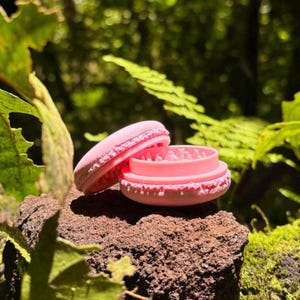 Peut inclure: Un moulin à herbes rose en forme de macaron, doté d'un bord extérieur texturé et d'une section supérieure ouverte révélant des dents de broyage internes. L'objet est composé de deux pièces circulaires, le couvercle supérieur étant incliné.