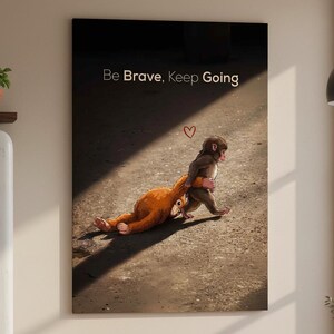 Op de afbeelding: Een canvas print met een illustratie van twee apen. De ene aap trekt de andere, met de tekst "Be Brave, Keep Going" en een hartsymbool. Het kunstwerk staat tegen een donkere achtergrond met een lichtstraal.