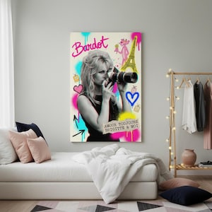Może przedstawiać: Wydruk na pł&oacute;tnie przedstawiający czarno-biały obraz Brigitte Bardot trzymającej aparat fotograficzny, z kolorowymi akcentami w stylu graffiti. Grafika zawiera r&oacute;żowe słowo "Bardot", r&oacute;żową panterę i wieżę Eiffla.