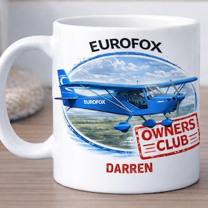 May include: White ceramic mug featuring a blue airplane design with the text "EUROFOX" and "OWNERS CLUB." The name "Darren" is printed in red. The mug has a white handle and is set against a blurred background.
