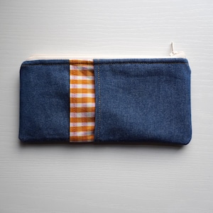 May include: A rectangular zippered pouch made of dark blue denim fabric. The front features a vertical accent strip of orange and white gingham check fabric. The pouch has a white zipper closure along the top edge and visible stitching details.