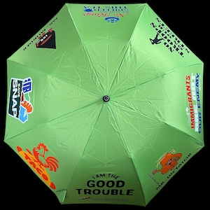 May include: A lime green umbrella with various graphics and text. The umbrella features phrases like "I AM THE GOOD TROUBLE" and "NO TROOPS ON OUR STREETS." Other graphics include a rooster, a cartoon figure, and the words "MR. MURDER."