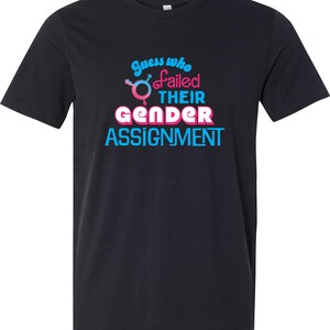 Könnte beinhalten: Ein schwarzes Kurzarm-T-Shirt mit dem Text "Guess who failed THEIR GENDER ASSIGNMENT" in blauer und rosa Schrift. Das Design enthält ein rosa-blaues Geschlechtssymbol-Icon, das in den Text auf der Vorderseite des T-Shirts integriert ist.