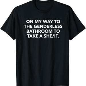 May include: A black short-sleeved t-shirt featuring white text printed on the chest that reads: ON MY WAY TO THE GENDERLESS BATHROOM TO TAKE A SHE/IT. The garment has a crew neckline and a standard fit, showcasing clear, bold lettering against the dark fabric.