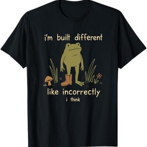 Pode incluir: Uma t-shirt preta de manga curta com um gráfico de um sapo verde a usar uma única bota castanha. O texto i'm built different está escrito acima do sapo e like incorrectly i think abaixo, acompanhado por um pequeno cogumelo e uma flor.