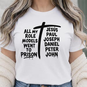 May include: White t-shirt with a black cross graphic and the text "ALL MY ROLE MODELS WENT TO PRISON" on the left side. On the right side, the text reads "JESUS PAUL JOSEPH DANIEL PETER JOHN".