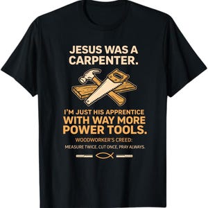 Op de afbeelding: Een zwart t-shirt met korte mouwen en een afbeelding van een hamer en zaag op een houten plank. De tekst luidt: JESUS WAS A CARPENTER. I'M JUST HIS APPRENTICE WITH WAY MORE POWER TOOLS. WOODWORKER'S CREED: MEASURE TWICE, CUT ONCE, PRAY ALWAYS.