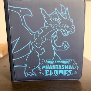 May include: A dark blue card binder featuring a neon blue outline of a dragon. The text "MEGA EVOLUTION PHANTASMAL FLAMES" is displayed in neon blue. The binder is standing upright on a dark brown surface.