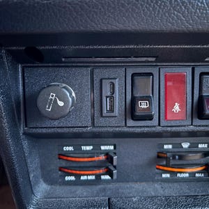 Può includere: Primo piano di un cruscotto nero per auto con vari comandi. Include un accendisigari, una porta USB, un pulsante delle luci di emergenza e cursori di controllo del clima con le etichette "Cool", "Temp" e "Warm". È visibile anche il controllo della miscela d'aria.