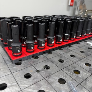 May include: A collection of black sockets arranged in a red organizer. The sockets are labeled with white numbers. The organizer is on a metal surface with evenly spaced holes.