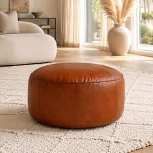 Pode incluir: Um pufe redondo de perfil baixo estofado em couro liso na cor conhaque. A peça apresenta detalhes de costura vertical visíveis ao longo das laterais e um painel superior circular, exibindo uma forma cilíndrica estruturada com acabamento uniforme.