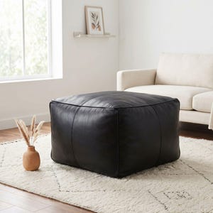 May include: A large, square black leather ottoman. The ottoman sits on a white rug with a geometric pattern. A beige sofa and a small vase with dried plants are in the background. The ottoman is a stylish piece of furniture.