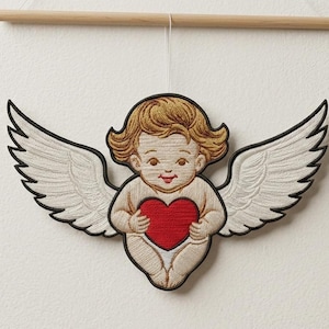 Puede incluir: Querubín bordado con alas sosteniendo un corazón rojo. El querubín tiene cabello rubio y está delineado en negro. Las alas son blancas con contornos negros. La obra de arte está suspendida de una clavija de madera.