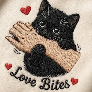 May include: Embroidered design featuring a black kitten playfully biting a hand. The design includes red hearts and the text "Love Bites" in a cursive font, all on a cream-colored fabric.