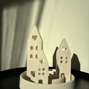 May include: A cream-colored ceramic cityscape sculpture featuring three stylized buildings of varying heights. Each building has cut-out windows and a heart-shaped opening. The sculpture sits on a circular base.