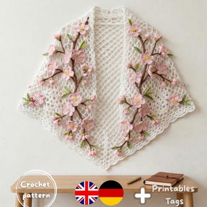 Puede incluir: Un chal blanco de ganchillo con flores rosas. El chal presenta flores rosas, hojas verdes y tallos marrones. El patrón de ganchillo es visible. El texto "Crochet pattern" y las banderas del Reino Unido y Alemania son visibles.