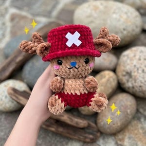 May include: A handmade plush toy of a cartoon character, featuring a red hat with a white cross, brown antlers, and a tan and red body. The toy has a blue nose and pink cheeks. The plush is held in a hand.