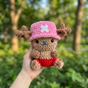 May include: A crochet plush toy featuring a tan creature with a pink hat, a white cross emblem, brown antlers, a blue nose, and red shorts. The plush is crafted from soft, textured yarn with black eyes and pink cheeks, showcasing a detailed design.