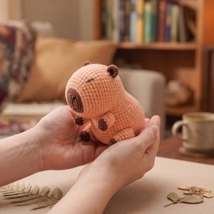 May include: A peach and brown crochet capybara toy. The handmade plush features closed eyes, small ears, and tiny feet. The capybara is held in open hands, highlighting the soft texture and detailed craftsmanship. The toy is a charming, handcrafted item.