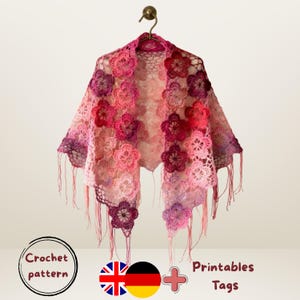 Può includere: Uno scialle all'uncinetto fatto a mano, in tonalità rosa e viola, decorato con applicazioni floreali. Lo scialle è esposto su una gruccia, mostrando il suo design intricato e i bordi sfrangiati. Le parole "Crochet pattern" sono visibili.